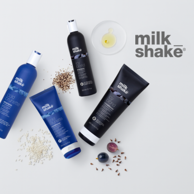milk_shake® Haarpflegeprodukte auf weißem Hintergrund, darunter zwei blaue Flaschen und eine Tube der „cold brunette“-Serie sowie zwei schwarze Verpackungen der „silver shine“-Linie. Umgeben von natürlichen Zutaten wie Reiskörnern, Leinsamen, Heidelbeeren, Weintrauben und Nelken. Oben rechts das milk_shake® Logo
