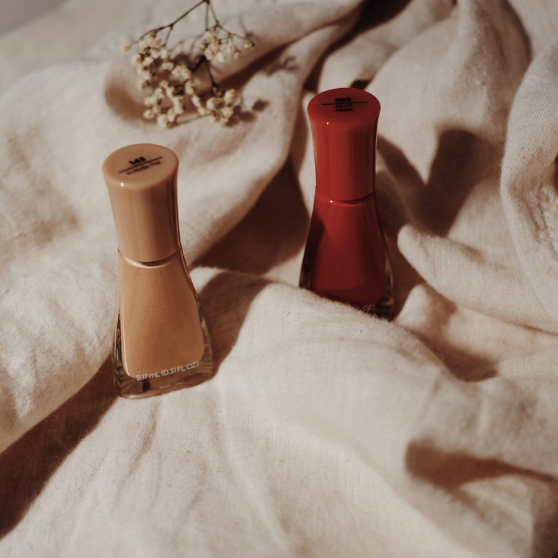 Zwei Nagellackflaschen in Beige und Rot auf beigefarbenem Stoff, dekoriert mit kleinen Trockenblüten.